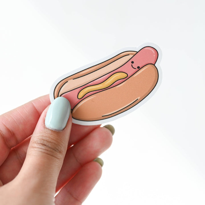 A hand holding a Hot Dog Sticker, a white sticker with an image of a smiling hot dog darnished with mustard.