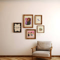 Five framed art prints in varying wood frames are arranged on a beige wall above a modern armchair with a light gray cushion and wooden frame.