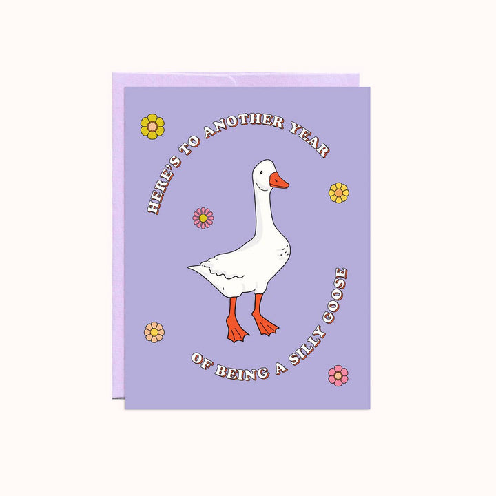 A purple greeting card with a white goose standing in the center. The goose has orange feet and a beak. The text 