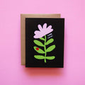 Good Luck Ladybug Encouragement Card. A simple, playful design featuring a large pink flower with green leaves on a black background. A red ladybug is perched on one of the leaves, and the text 'Good Luck!' is written in white next to the flower. A kraft paper envelope is partially visible behind the card, adding a rustic touch.