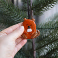 A hand holds a small orange-brown frog ornament with a black hanging ribbon, set against green pine tree branches.