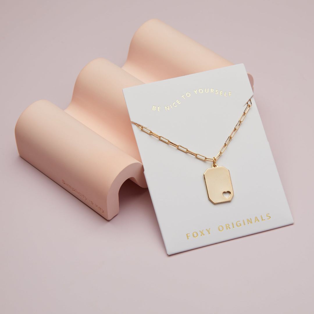 Gold necklace with a tag on a 'Foxy Originals' card against a pink background