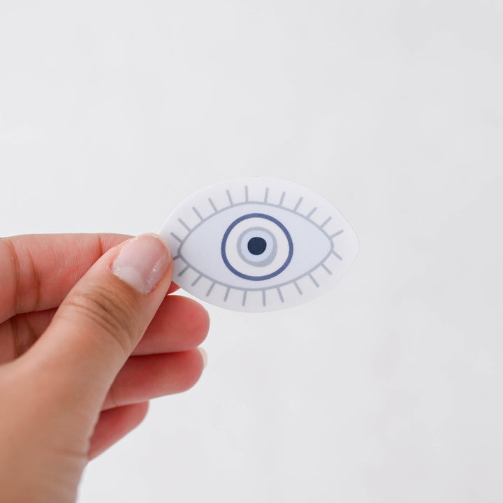 A hand holding a Evil Eye Sticker, a white sticker with a blue evil eye.