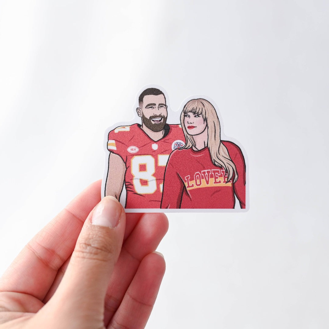 A hand holding a Taylor and Travis Sticker, a white sticker with an image of Travis Kelce in his football jersey and Taylor Swift in a Chiefs colours sweater that reads LOVER.