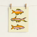 A vertical art print featuring three stylized fish illustrations in orange, pink, and yellow-green tones on a cream background, hanging from two wooden clips on a white wall.