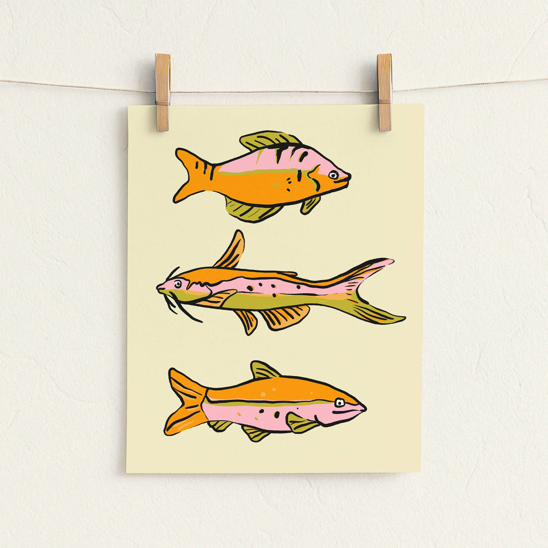 A vertical art print featuring three stylized fish illustrations in orange, pink, and yellow-green tones on a cream background, hanging from two wooden clips on a white wall.