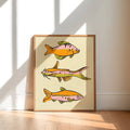 A framed art print featuring three stylized orange and pink fish on a cream background, leaning against a white wall on a light wood floor with natural window shadows.