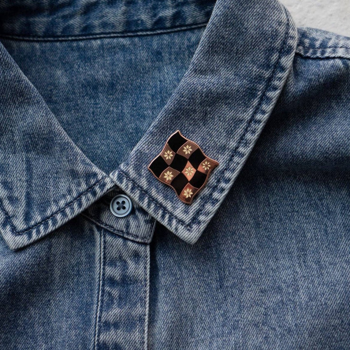 Rose gold-toned pin shaped like a checkered flag with black and white squares. Each square has a small, white flower design in the center.
