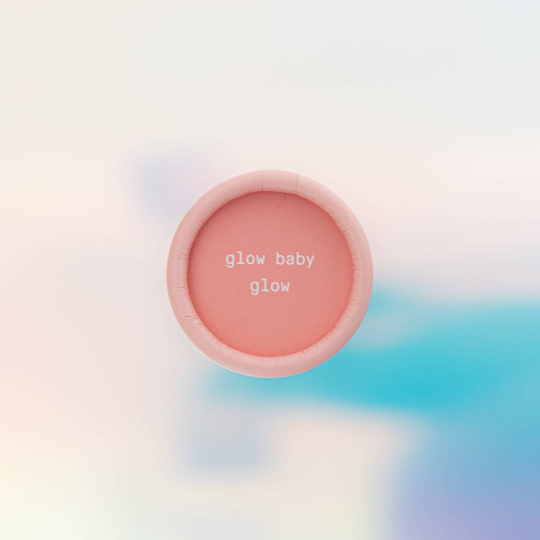 Sunset Glow Stick, a solid highlighter makeup stick in a warm coral pink sunset colour. It is presented in a cardboard tube, with a pull off top.