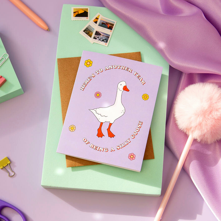 A purple greeting card with a white goose standing in the center. The goose has orange feet and a beak. The text 