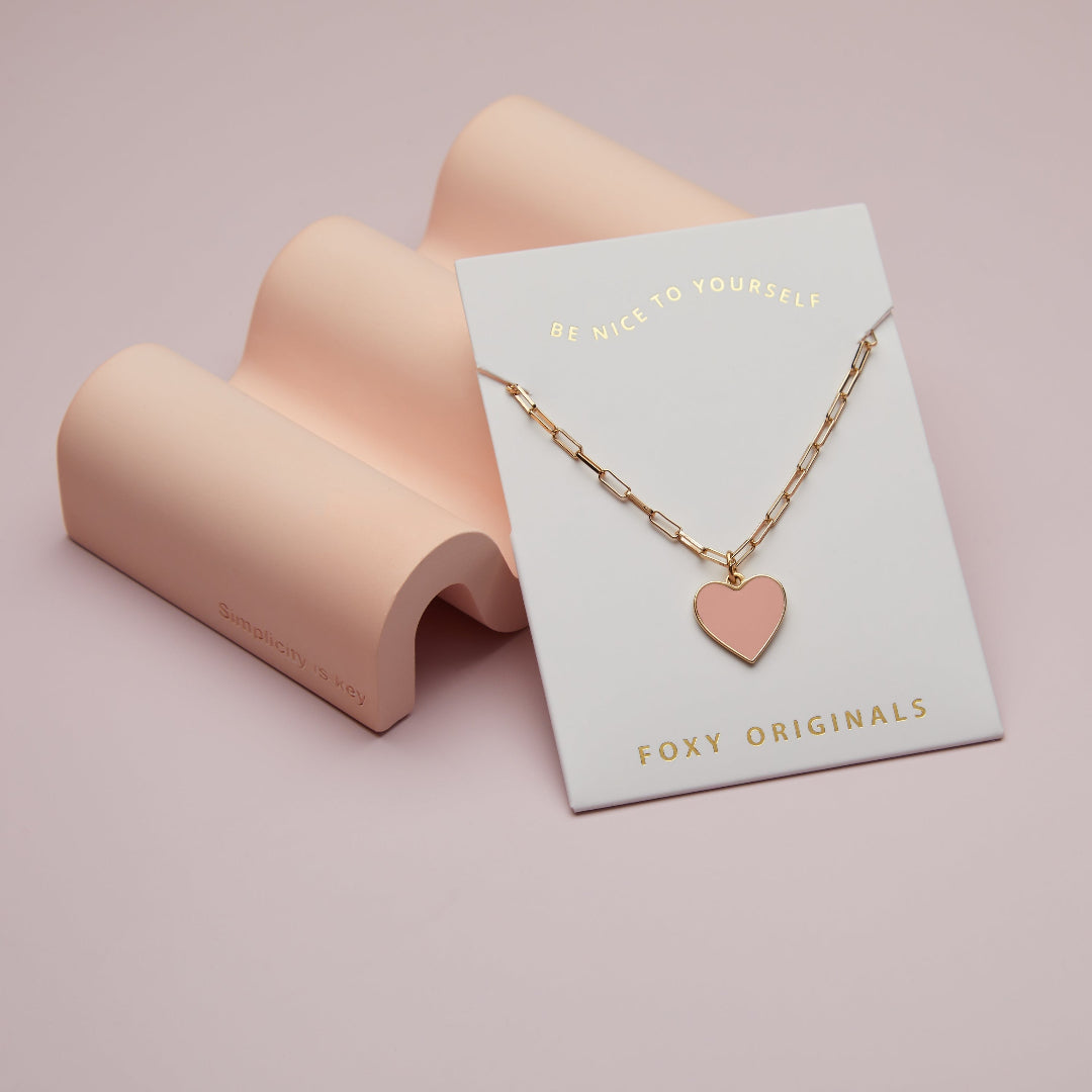 Gold necklace with a heart pendant on a 'Foxy Originals' card against a pink background
