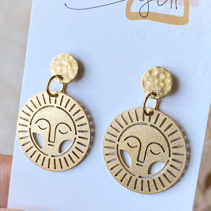 Brass Viviette Earrings. These elegant earrings feature circular brass discs with a sunburst cutout design and a small hammered brass circle at the top. The earrings are displayed on a soft, neutral-toned surface, adding a warm and sophisticated touch.
