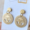 Brass Viviette Earrings. These elegant earrings feature circular brass discs with a sunburst cutout design and a small hammered brass circle at the top. The earrings are displayed on a soft, neutral-toned surface, adding a warm and sophisticated touch.