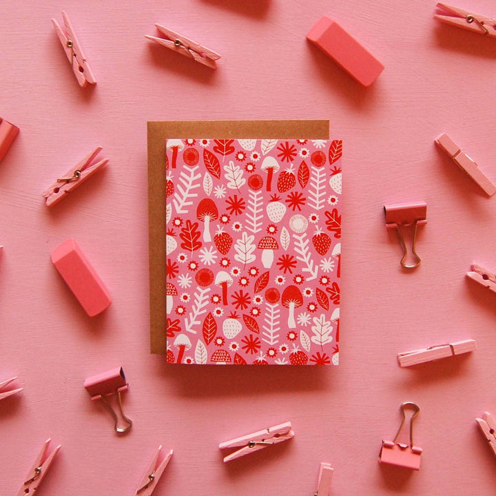 Woodlands-themed greeting card titled ‘Woodlands Card,’ with a pink background adorned by white, red, and orange illustrations of mushrooms, leaves, acorns, and flowers.