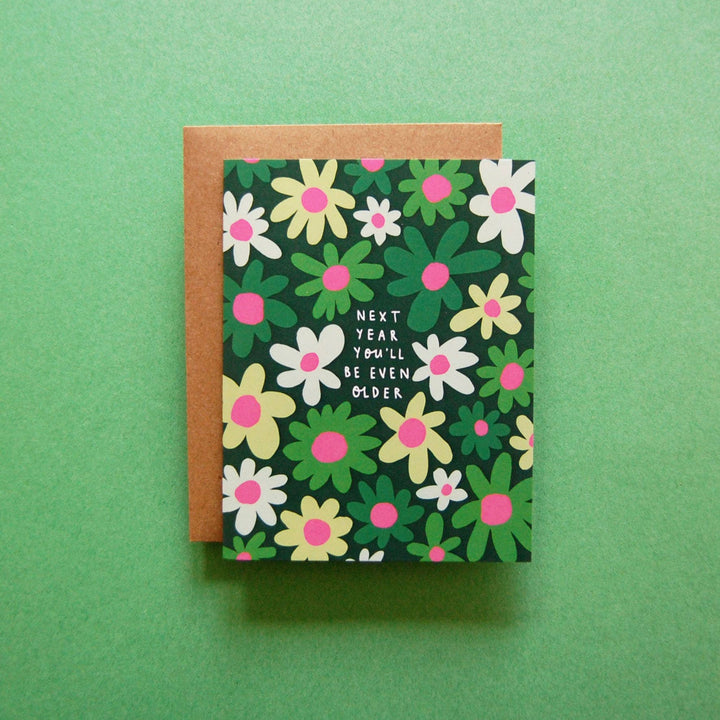 Next Year Birthday Card: A humorous birthday card featuring a dark green background with stylized white, light green, and pink flowers. In the center, white text reads ‘NEXT YEAR YOU’LL BE EVEN OLDER.’