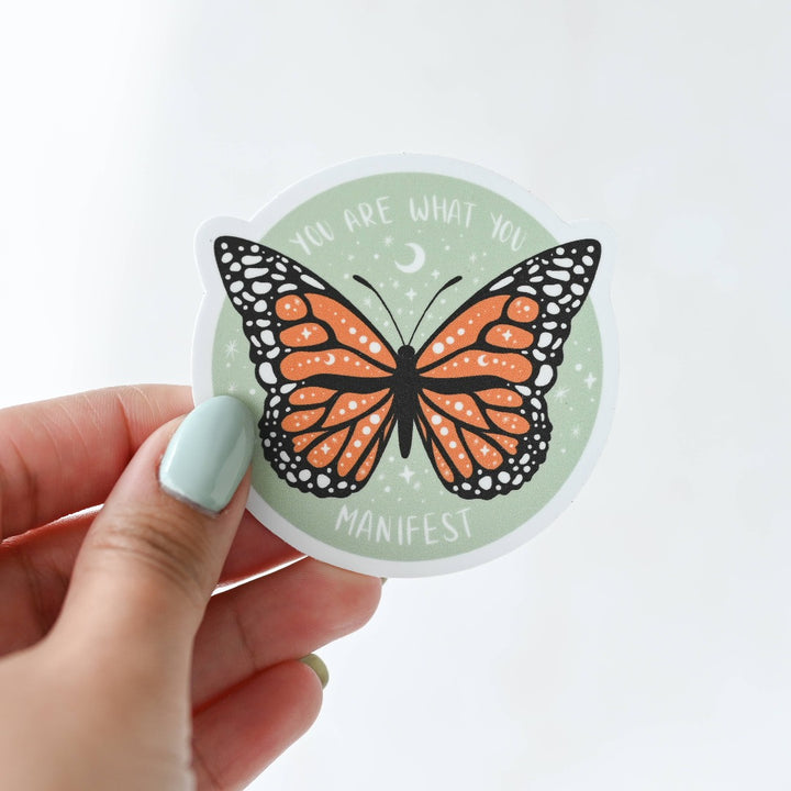 A hand holding You Are What You Manifest Sticker, a green sticker with a monarch butterfly in the middle with the words 