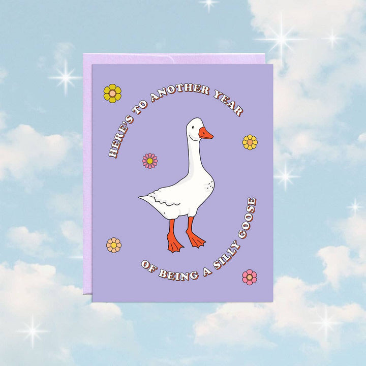 A purple greeting card with a white goose standing in the center. The goose has orange feet and a beak. The text 