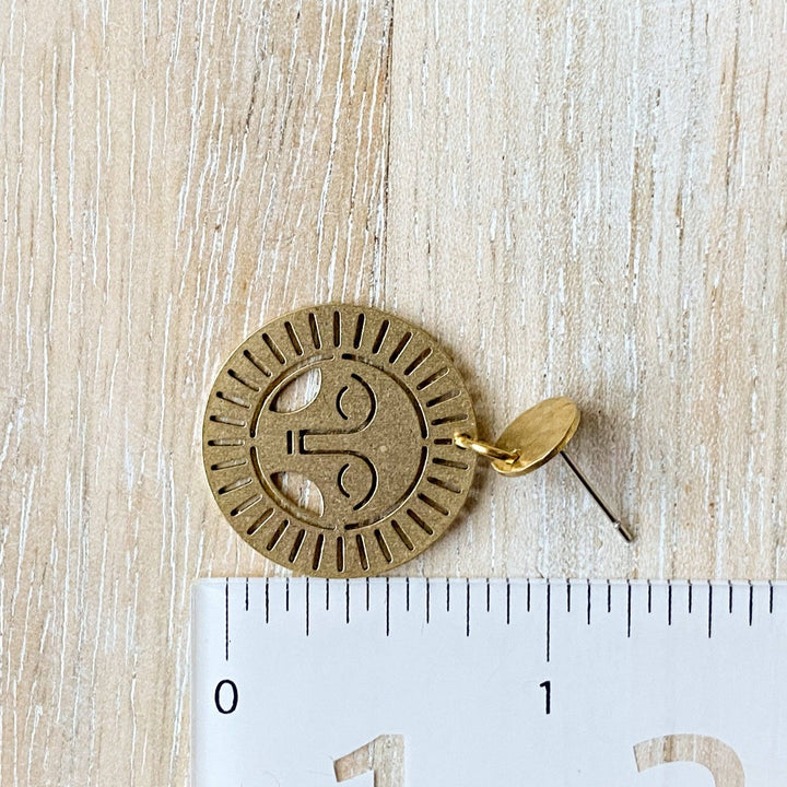 Brass Viviette Earrings. These elegant earrings feature circular brass discs with a sunburst cutout design and a small hammered brass circle at the top. The earrings are displayed on a soft, neutral-toned surface, adding a warm and sophisticated touch.