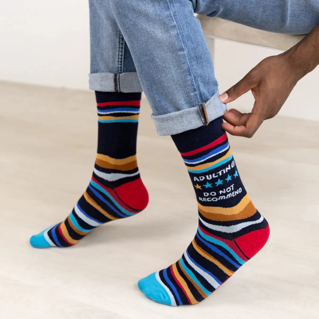 A pair of crew socks with a humorous message. The socks are mostly black with colorful stripes. The text "Adulting" is printed at the top, followed by a star rating of three stars and the phrase "Do Not Recommend."