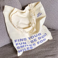 A white tote bag with blue text. The text reads "FIND YOUR FUN & BE BOLD SMILES FOR MILES" with a smiley face symbol between each phrase. The bottom right corner displays the text "epic blend."
