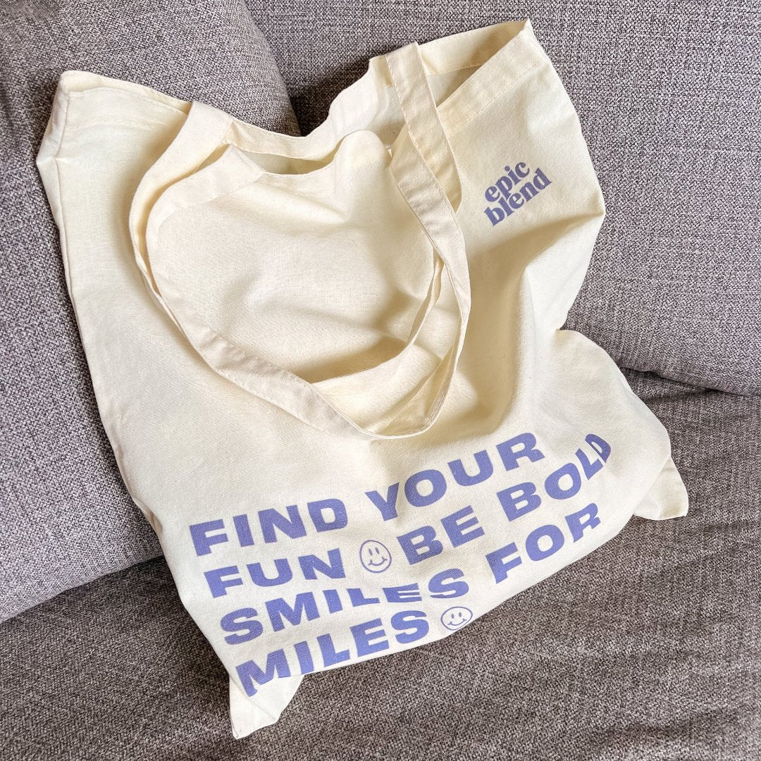 A white tote bag with blue text. The text reads "FIND YOUR FUN & BE BOLD SMILES FOR MILES" with a smiley face symbol between each phrase. The bottom right corner displays the text "epic blend."