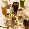 A styled flat-lay of multiple MY CAFE dropper bottles and prepared drinks on a warm cream surface. Visible products include several MY CAFE bottles with gold caps, a mug of iced coffee with whipped cream and a straw, a tall green drink, a black mug with a hot drink, a bowl of pink drink, a cup of green matcha, and a latte art coffee mug.