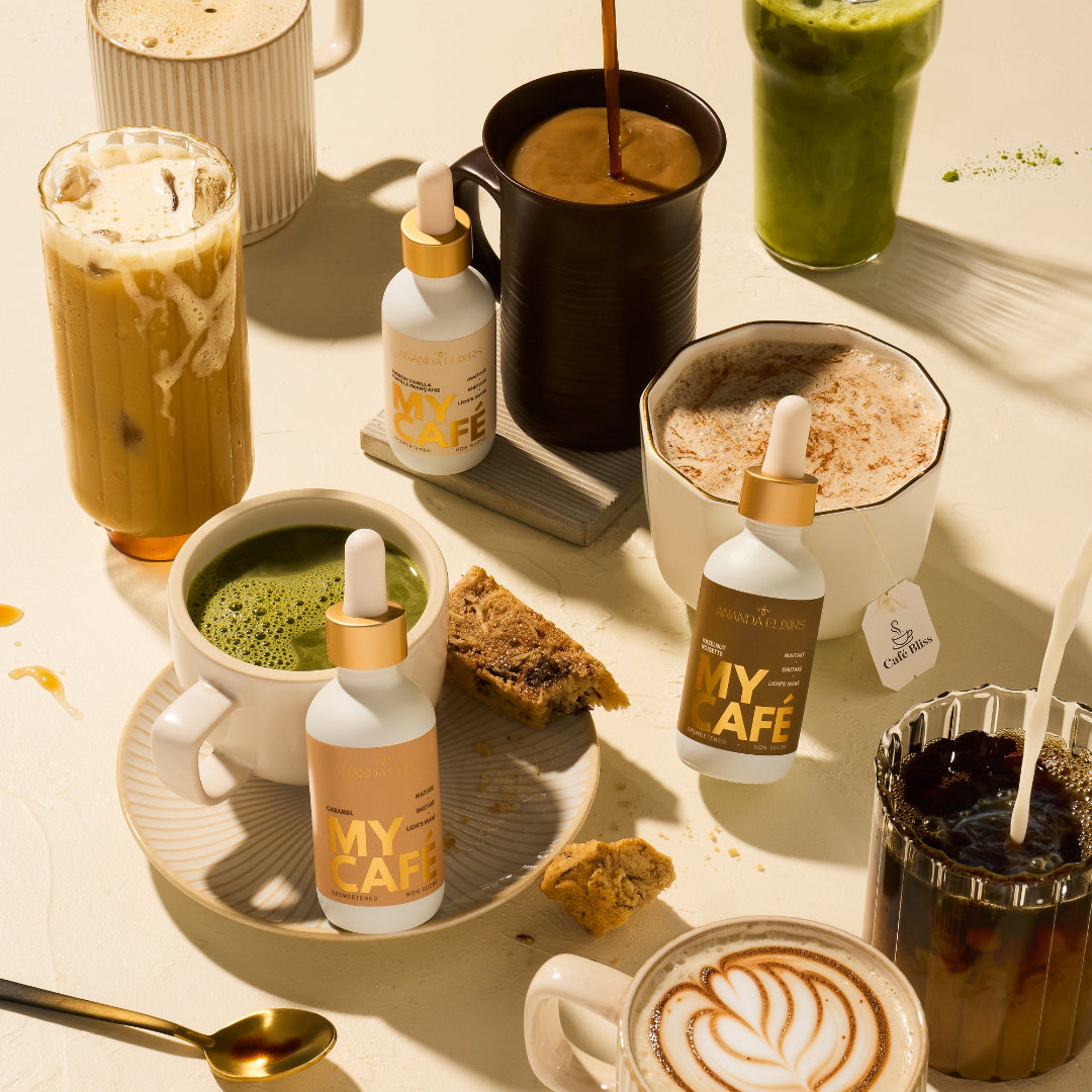 A styled flat-lay of multiple MY CAFE dropper bottles and prepared drinks on a warm cream surface. Visible products include several MY CAFE bottles with gold caps, a mug of iced coffee with whipped cream and a straw, a tall green drink, a black mug with a hot drink, a bowl of pink drink, a cup of green matcha, and a latte art coffee mug.