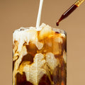 An iced coffee or cola drink in a clear glass with ice, layered with creamy white milk and dark brown coffee, shown against a warm beige background with a dropper above.
