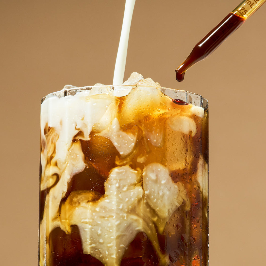 An iced coffee or cola drink in a clear glass with ice, layered with creamy white milk and dark brown coffee, shown against a warm beige background with a dropper above.