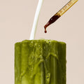 A close-up of a frosted green glass candle with white wax dripping down the sides, topped with a white stick, while a glass dropper adds a dark brown liquid against a solid beige background.