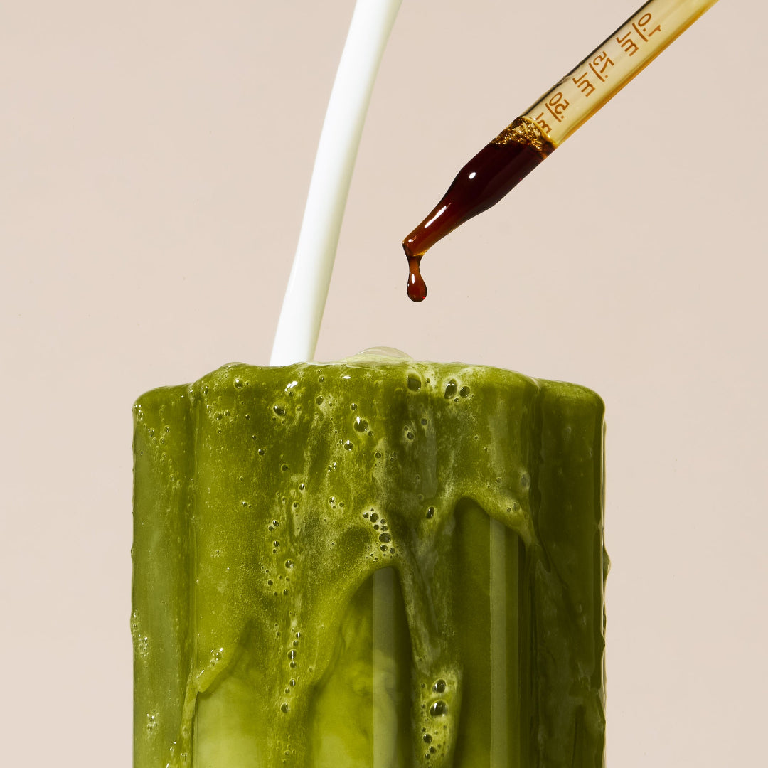 A close-up of a frosted green glass candle with white wax dripping down the sides, topped with a white stick, while a glass dropper adds a dark brown liquid against a solid beige background.