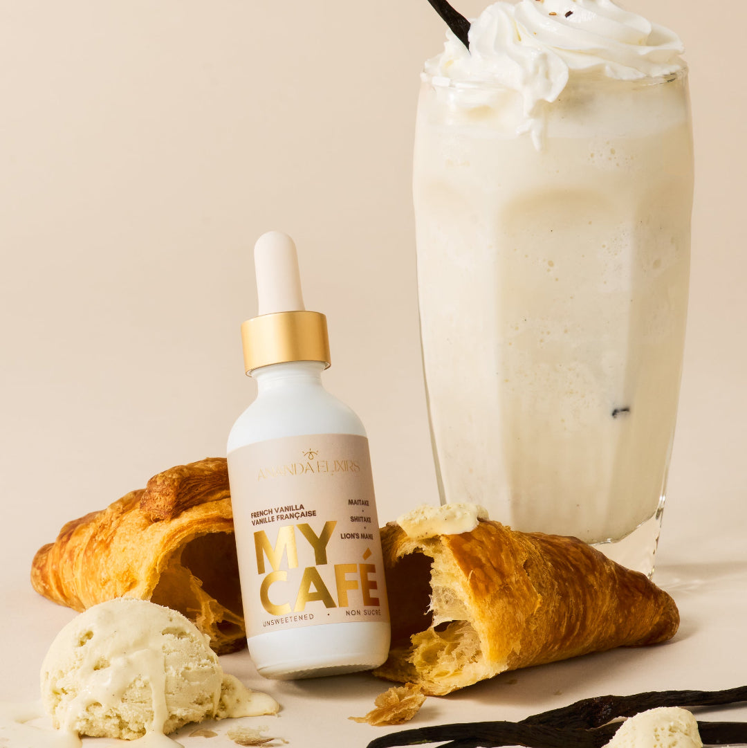 A small white dropper bottle labeled 'MY CAFE' with a gold collar stands beside a tall glass of creamy vanilla milkshake topped with whipped cream and a drizzle of syrup. Croissant pieces, vanilla ice cream scoops, vanilla beans, and a black straw are arranged on a warm beige background.