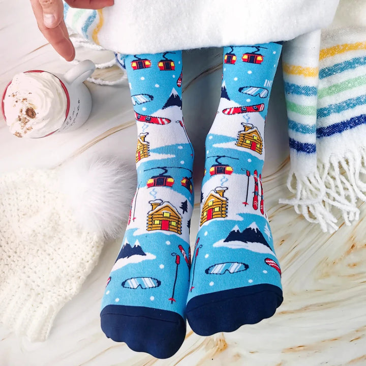 A pair of blue socks with a winter sports theme. The socks feature illustrations of skiers, snowboards, and a mountain cabin. The text 