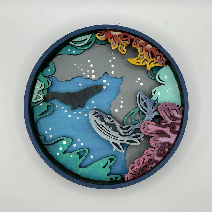 A Paint By Layers Craft Kit featuring a circular shadow box with a pre-designed underwater scene. The scene depicts whales swimming in the ocean, surrounded by coral reefs and bubbles. The kit includes layers of wood, paint, and instructions. The title of the kit is 