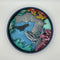 A Paint By Layers Craft Kit featuring a circular shadow box with a pre-designed underwater scene. The scene depicts whales swimming in the ocean, surrounded by coral reefs and bubbles. The kit includes layers of wood, paint, and instructions. The title of the kit is 