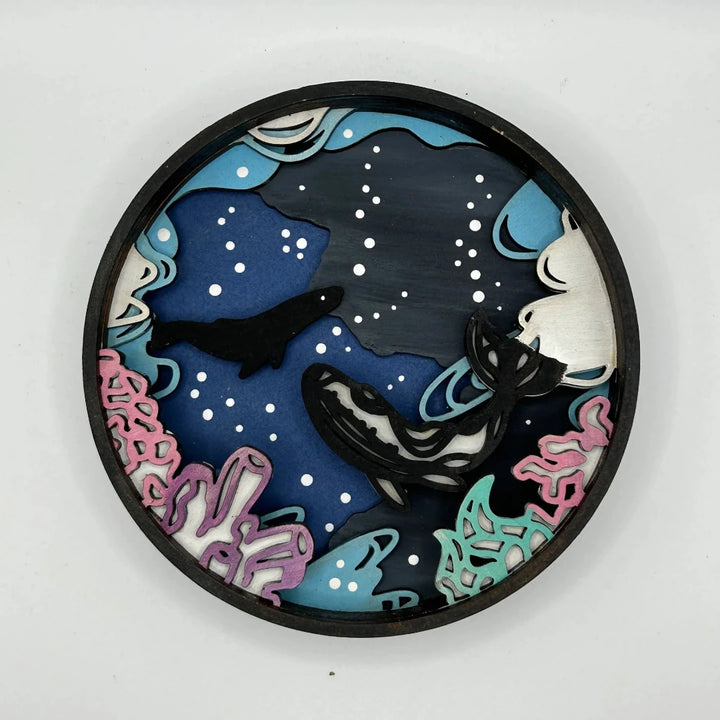 A Paint By Layers Craft Kit featuring a circular shadow box with a pre-designed underwater scene. The scene depicts whales swimming in the ocean, surrounded by coral reefs and bubbles. The kit includes layers of wood, paint, and instructions. The title of the kit is 