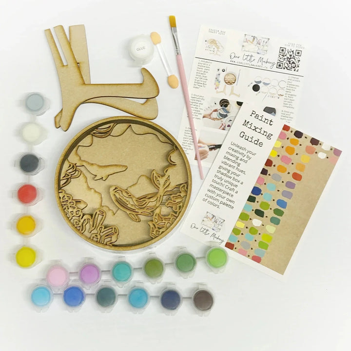 A Paint By Layers Craft Kit featuring a circular shadow box with a pre-designed underwater scene. The scene depicts whales swimming in the ocean, surrounded by coral reefs and bubbles. The kit includes layers of wood, paint, and instructions. The title of the kit is 