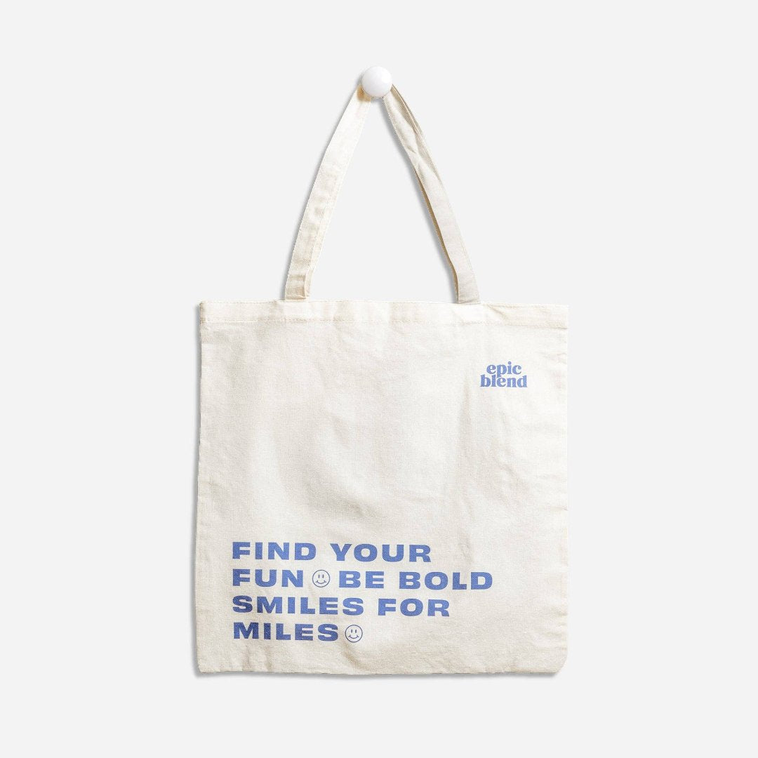 A white tote bag with blue text. The text reads "FIND YOUR FUN & BE BOLD SMILES FOR MILES" with a smiley face symbol between each phrase. The bottom right corner displays the text "epic blend."