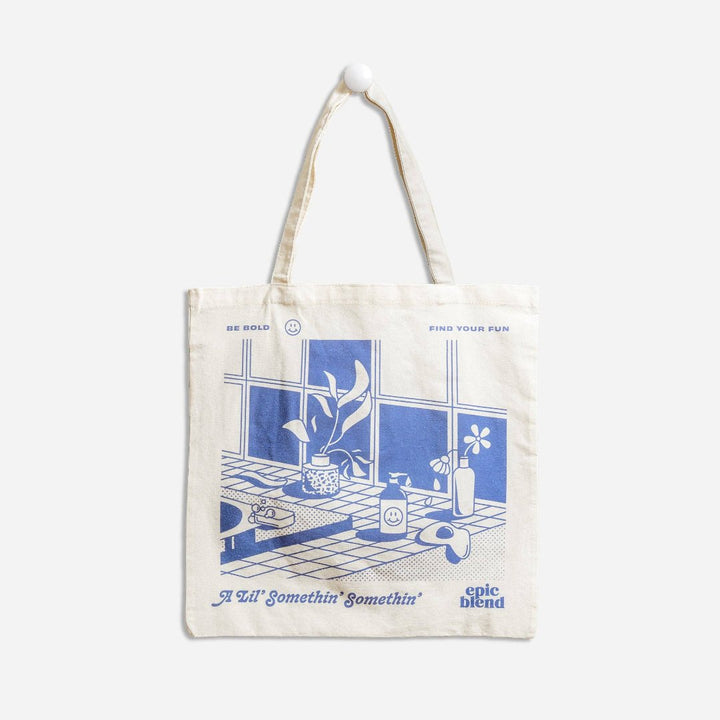 A white tote bag with a blue and white illustration. The illustration shows a plant on a windowsill, with a sink, soap, and other bathroom items visible. The text on the bag includes 
