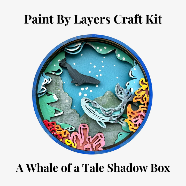 A Paint By Layers Craft Kit featuring a circular shadow box with a pre-designed underwater scene. The scene depicts whales swimming in the ocean, surrounded by coral reefs and bubbles. The kit includes layers of wood, paint, and instructions. The title of the kit is 