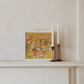 Framed artwork with flowers on a shelf next to two white candles in bronze candle holders.
