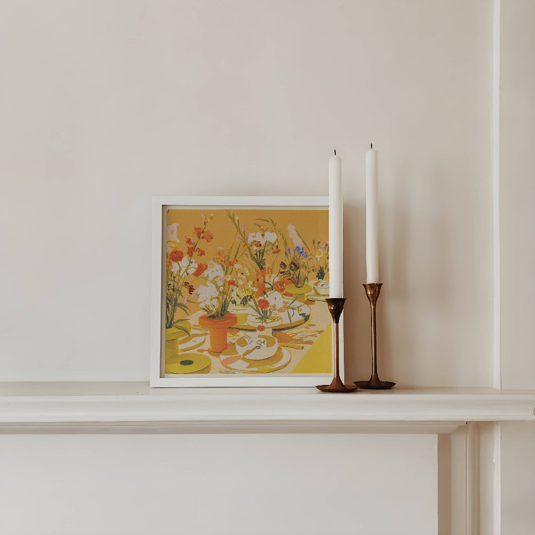 Framed artwork with flowers on a shelf next to two white candles in bronze candle holders.