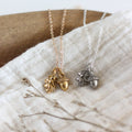 Two acorn-shaped necklaces, one gold and one silver, on a textured fabric background.