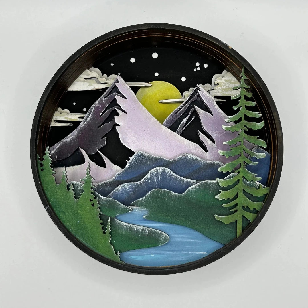 A Paint By Layers Craft Kit featuring a circular shadow box with a pre-designed landscape scene. The scene depicts a mountain range under a starry night sky, with a river flowing through a valley. The kit includes layers of wood, paint, and instructions. The title of the kit is "Adventure Awaits Shadow Box."