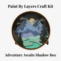 A Paint By Layers Craft Kit featuring a circular shadow box with a pre-designed landscape scene. The scene depicts a mountain range under a starry night sky, with a river flowing through a valley. The kit includes layers of wood, paint, and instructions. The title of the kit is "Adventure Awaits Shadow Box."
