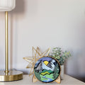 A round wooden decorative plaque featuring a colorful mountain landscape with a sun, trees, and a river, displayed on a tabletop next to a gold star decor piece and a potted plant.