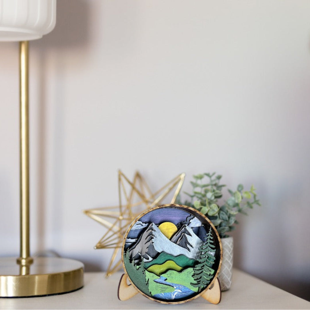 A round wooden decorative plaque featuring a colorful mountain landscape with a sun, trees, and a river, displayed on a tabletop next to a gold star decor piece and a potted plant.