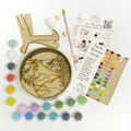 A Paint By Layers Craft Kit featuring a circular shadow box with a pre-designed landscape scene. The scene depicts a mountain range under a starry night sky, with a river flowing through a valley. The kit includes layers of wood, paint, and instructions. The title of the kit is "Adventure Awaits Shadow Box."