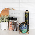 A styled kitchen countertop display featuring a black cylindrical tin of Nutra Organic Mountain Olive Oil, two glass spice grinders, a small round landscape art print on a wooden stand, a white book, and a potted plant against a white subway tile background.