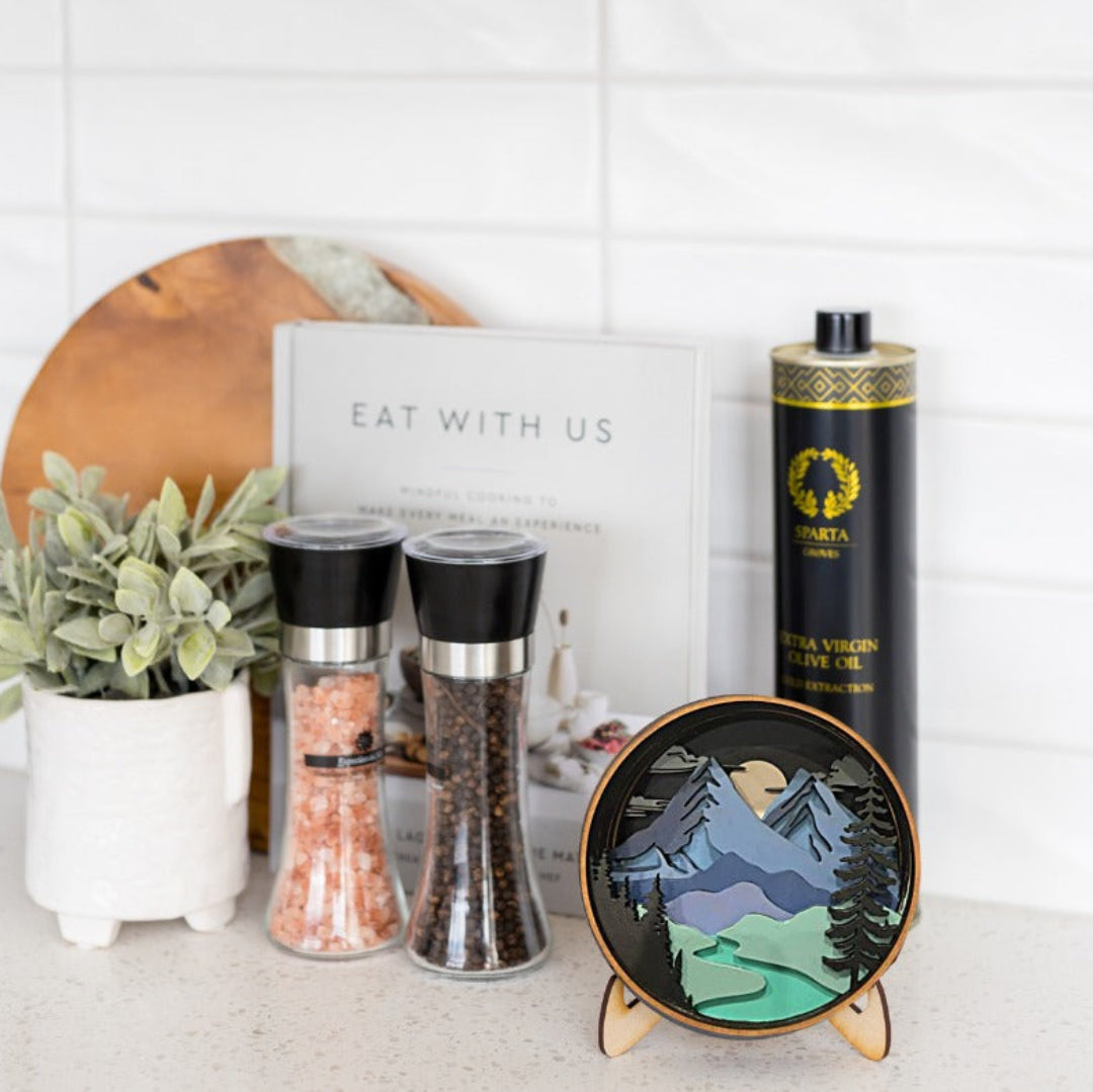 A styled kitchen countertop display featuring a black cylindrical tin of Nutra Organic Mountain Olive Oil, two glass spice grinders, a small round landscape art print on a wooden stand, a white book, and a potted plant against a white subway tile background.